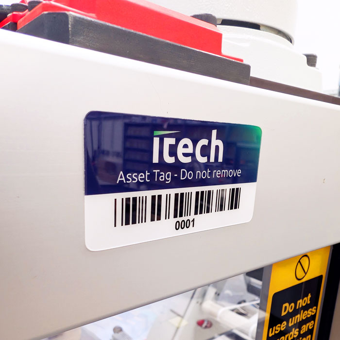 Close-up of an Itech asset tag with a barcode and the text 'Asset Tag - Do not remove' attached to light-colored equipment, showing serial 0001.